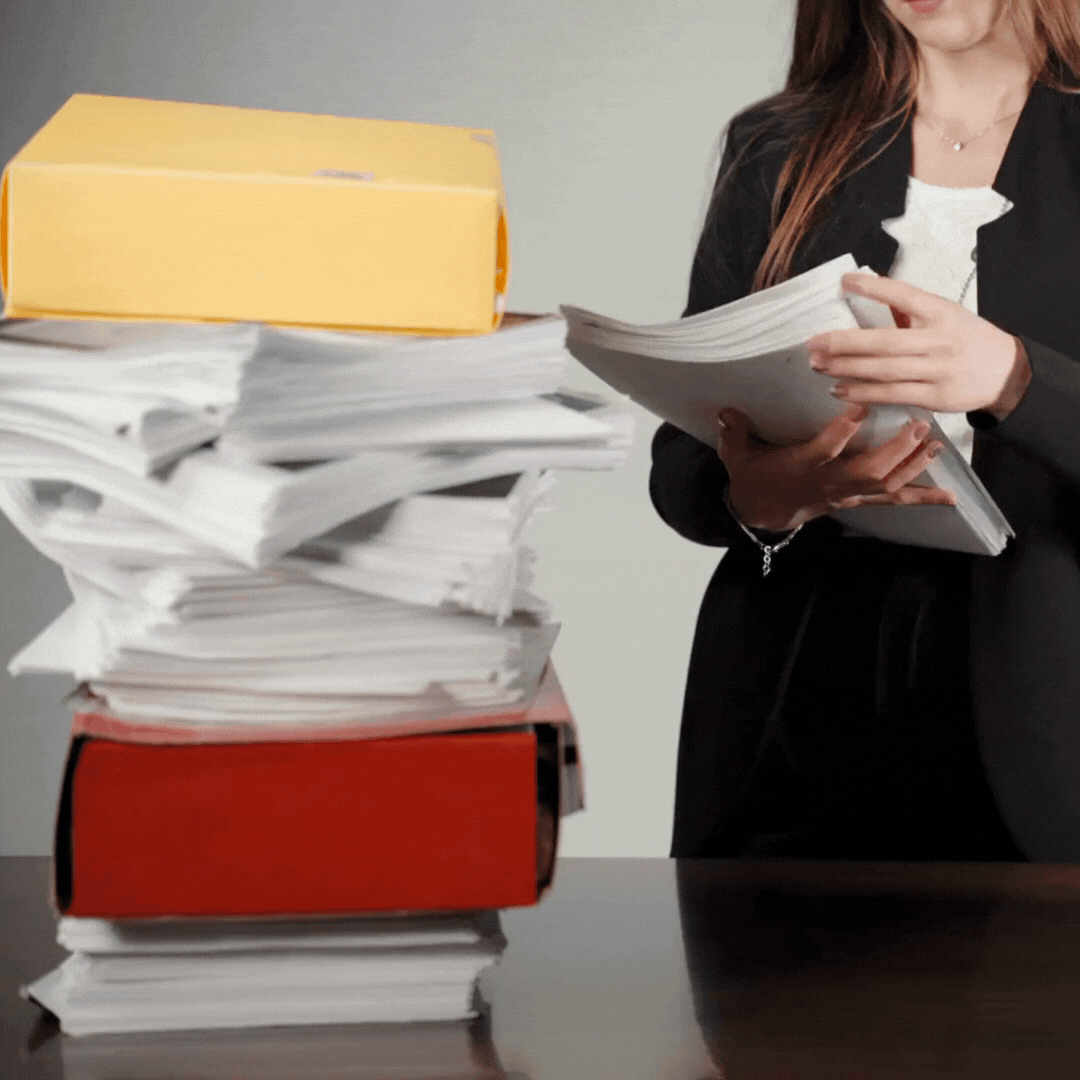 Professional in a black blazer reviewing documents with a tall stack of paperwork and colored file folders on the desk.