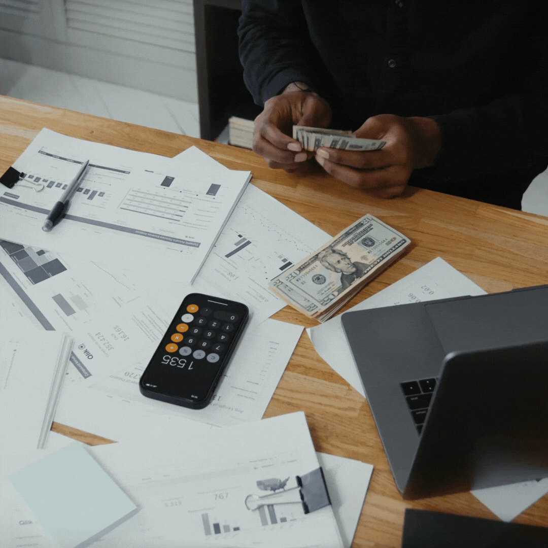 Person counting cash on a desk with financial documents, a calculator app, and a laptop, representing cost savings.